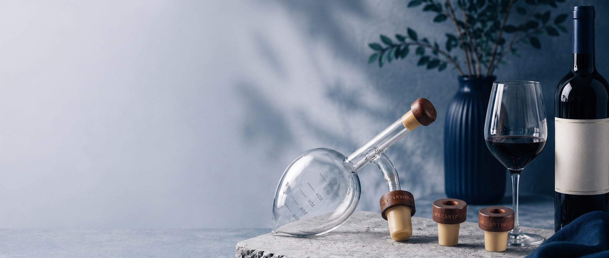Oxigenador Decantorino de cristal junto a una copa de vino tinto, una botella y tapones de madera